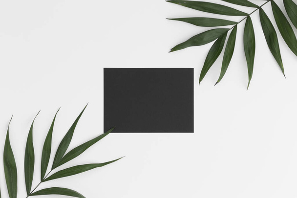 Vista superior de una maqueta de tarjeta negra con decoración de hojas de palma sobre una mesa blanca
. - Foto, imagen