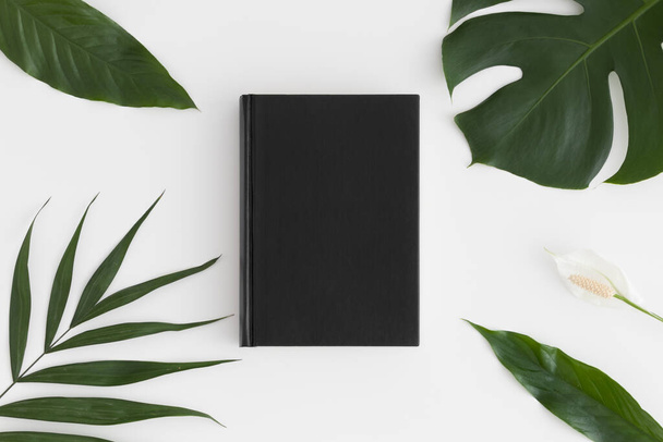 Top view of a black book mockup with tropical leaves decoration on a white table. - Photo, Image