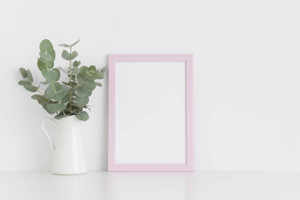 Pinkfarbene Rahmen-Attrappe mit einem Strauß Eukalyptus in einer Vase auf einem weißen Tisch.. - Foto, Bild