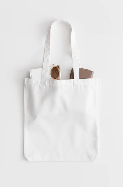 Λευκό tote τσάντα mockup με ένα βιβλίο, πορτοφόλι και γυαλιά σε ένα λευκό τραπέζι. - Φωτογραφία, εικόνα