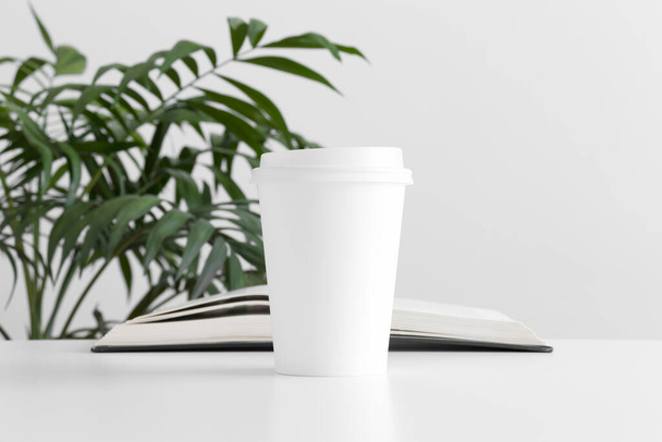Coffee paper cup mockup with a book and a palm plant on a white table. - Фото, изображение