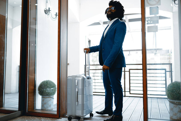 Hombre negro en traje formal con maleta empacada entrando en la puerta del hotel - Foto, Imagen