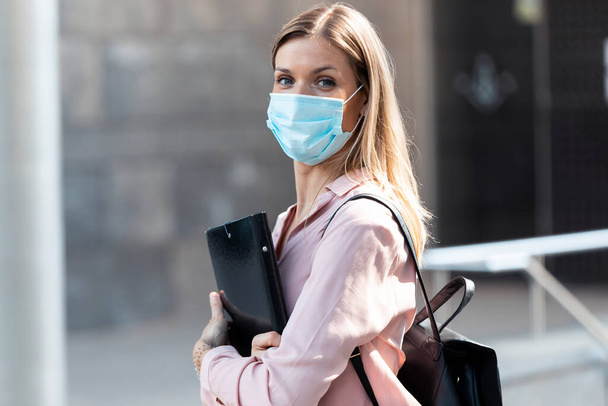 Shot of a beautiful blonde woman in face mask with a folder looking at camera while standing in the street. - Photo, Image