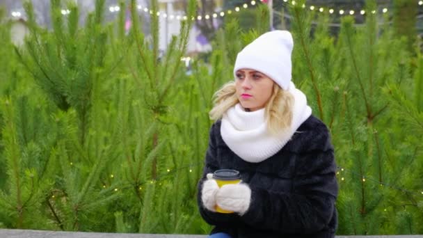 Γυναίκα σε ζεστό λευκό καπέλο, κασκόλ, γάντια κάθεται κρατώντας καφέ χάρτινο κύπελλο στα χέρια - Πλάνα, βίντεο