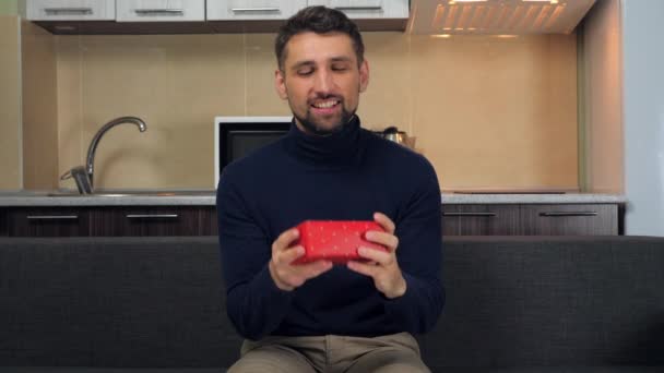 Glücklich schöner Mann im Pullover fängt rote Schachtel, untersucht, lächelt, freut sich Geschenk - Filmmaterial, Video