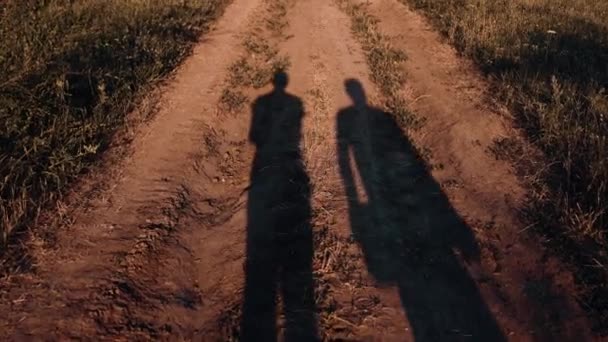 Der Schatten zweier Männer, verursacht durch das Sonnenlicht bei Sonnenuntergang, das sich auf einer Landstraße inmitten eines Feldes an einem Hang spiegelt - Filmmaterial, Video