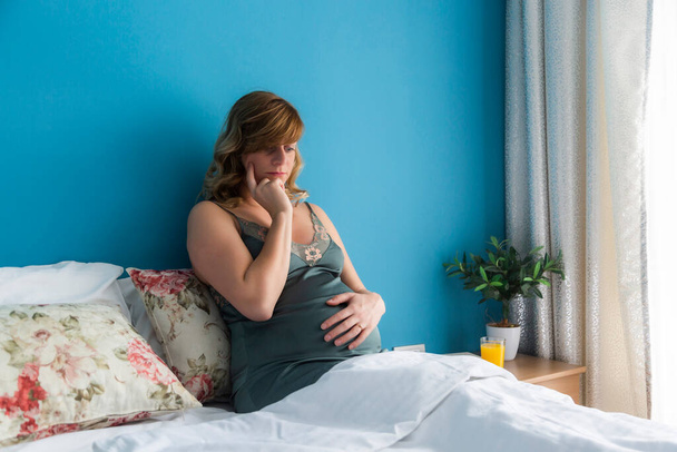 Een zwangere vrouw in een semi-zittende positie in bed met haar handen rond haar buik, zich afvragend over een baby. Ze ziet er attent uit.. - Foto, afbeelding