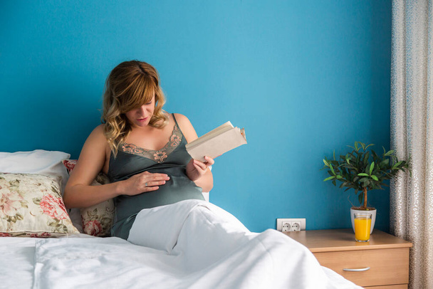 Egy terhes nő érzi a baba mozgását az ágyában, miközben könyvet olvas. Az arckifejezése azt mutatja, hogy a baba a gyomrában mozog.. - Fotó, kép