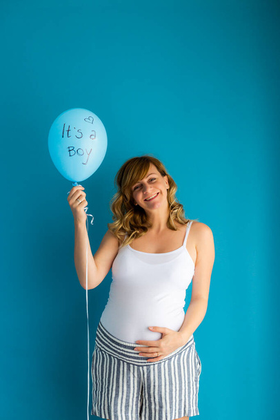 Беременная улыбающаяся женщина держит воздушный шар со словами "это мальчик". Она перед синей стеной.. - Фото, изображение
