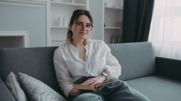 Young beautiful brunette with stylish glasses and white shirt sits on sofa in modern bright living room with book in hand. Portrait of happy, smiling student girl in close-up. Homework and education - Materiaali, video