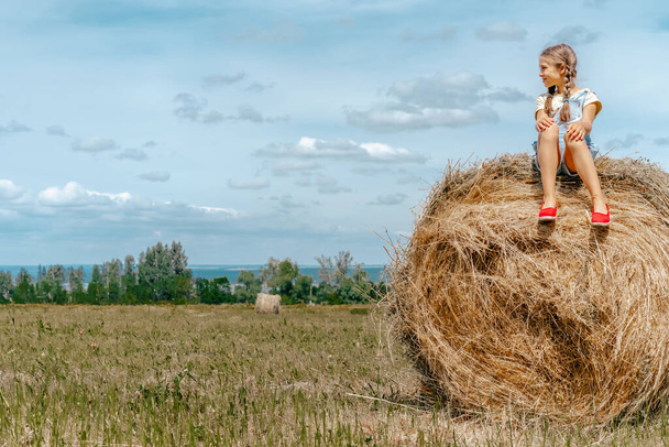  Niña niña en overoles de mezclilla sentada en fardos de paja, caminando en la naturaleza en el pueblo
 - Foto, imagen