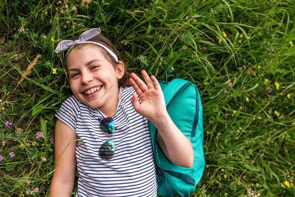 Милая маленькая девочка улыбается и машет привет, пока лежала с рюкзаком на траве летом - Фото, изображение