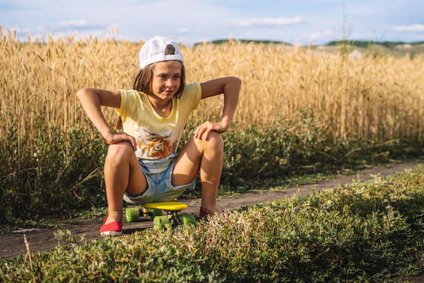 Mała dziewczynka w dżinsowym kombinezonie siedzi na deskorolce na środku pola pszenicy, bezczelny niegrzeczny nastolatek - Zdjęcie, obraz