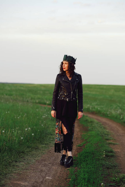 A young woman of 25-30 years old with curly red hair, in a black leather jacket and a tiara with feathers walks along a country road. - Foto, afbeelding