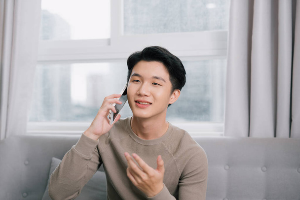 Atraktivní mladý muž mluví na mobilním telefonu a usmívá se, zatímco sedí na pohovce doma - Fotografie, Obrázek