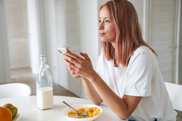 Photo of young caucasian woman with pink hair using cellphone while having breakfast in white kitchen - Fotó, kép