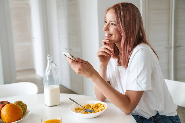 Photo of young cheerful woman with pink hair using cellphone while having breakfast in white kitchen - Φωτογραφία, εικόνα