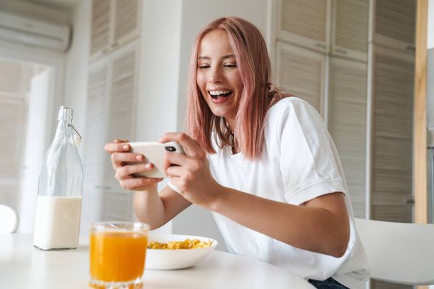 Photo of young excited woman playing online game on cellphone while having breakfast in white kitchen - Fotografie, Obrázek