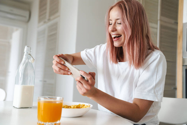 Photo of young excited woman playing online game on cellphone while having breakfast in white kitchen - Fotografie, Obrázek