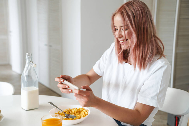 Photo of young excited woman playing online game on cellphone while having breakfast in white kitchen - Foto, afbeelding