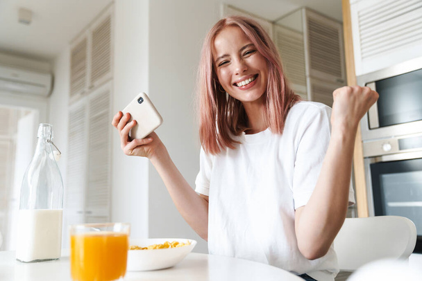 Foto di giovane donna eccitata fare gesto vincitore durante il gioco online sul cellulare in cucina bianca - Foto, immagini