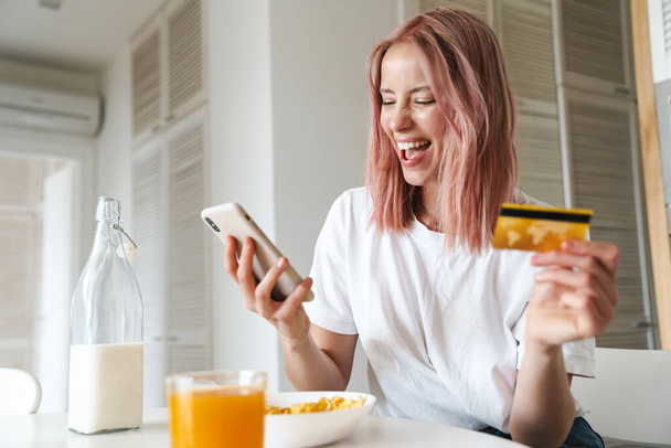 Photo of young laughing woman holding credit card and cellphone while having breakfast in white kitchen - Valokuva, kuva