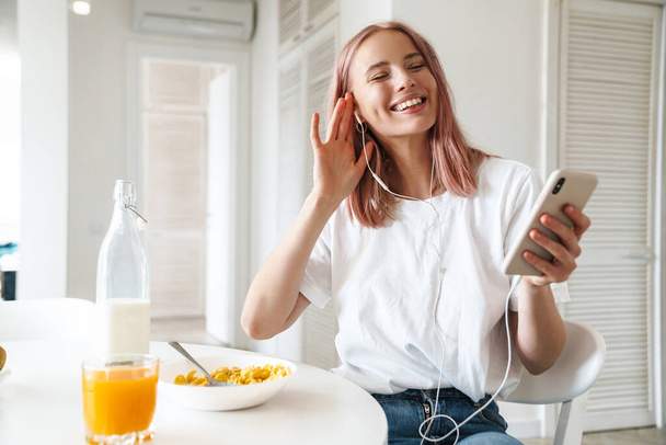 Photo of nice cheerful woman using cellphone and earphones while having breakfast in white kitchen - Foto, Imagen