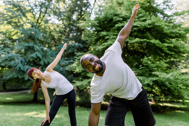 Beautiful multiethnic couple in sports clothes is stretching on yoga mats while working out outdoors at city park. Fitness and sport together. Focus on smiling handsome African man - Fotoğraf, Görsel