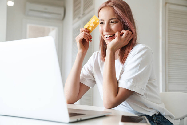 Photo of cheerful woman working with laptop and holding credit card while sitting at table in kitchen - Фото, зображення