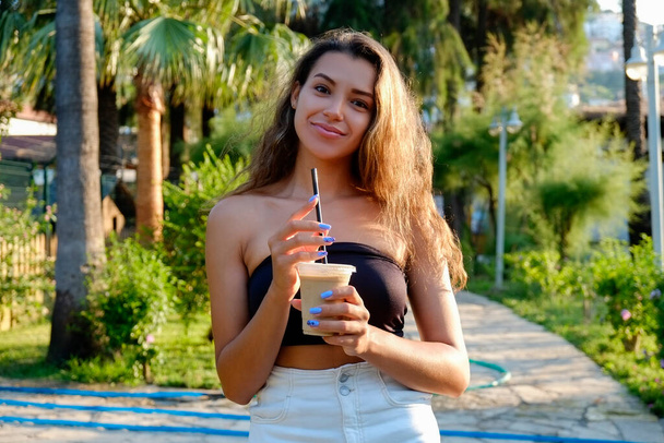 Portrait of young beautiful woman with long wavy brunette hair drinking iced coffee in tropical destination park with greenery on the background. Joy of summer holidays concept. Close up, copy space. - Foto, imagen