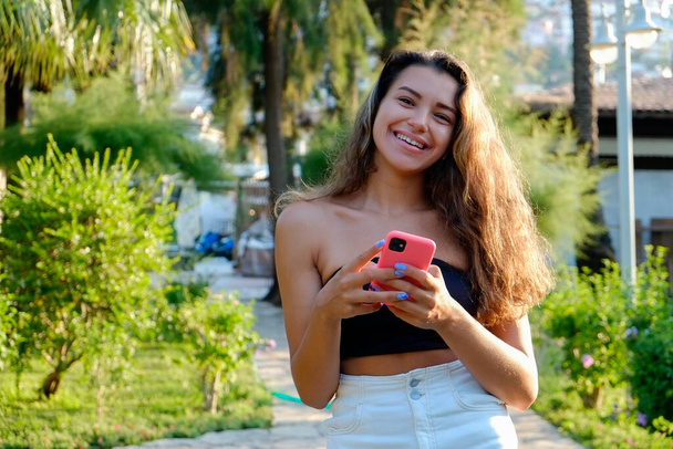 Portrait of young beautiful woman with long wavy brunette hair with her phone in tropical destination park with greenery on the background. Joy of summer holidays concept. Close up, copy space. - Photo, image