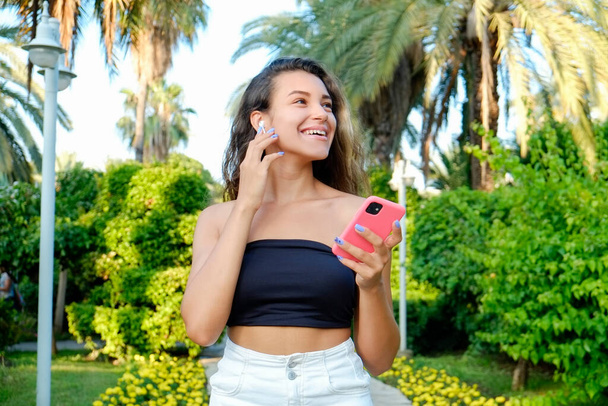 Portrait of young beautiful woman with long wavy brunette hair with her phone in tropical destination park with greenery on the background. Joy of summer holidays concept. Close up, copy space. - Photo, Image