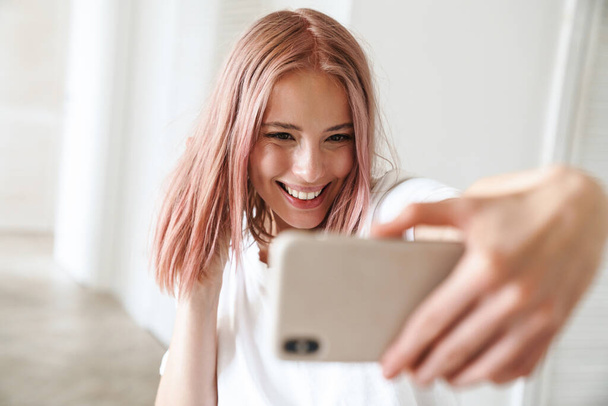 Photo of caucasian excited woman with pink hair smiling and taking selfie photo on mobile phone indoors - Fotografie, Obrázek