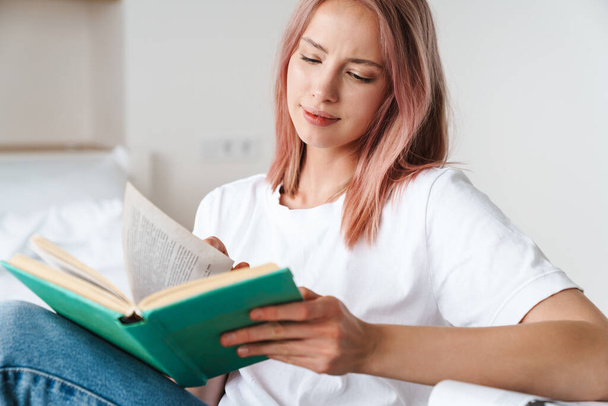 Image of focused pretty woman with pink hair reading book and thinking while resting at home - Foto, immagini