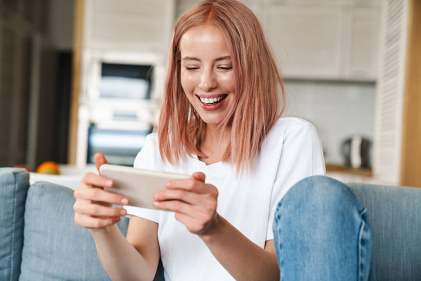 Image of delighted woman playing video game on mobile phone while sitting on sofa at home - Foto, Imagem