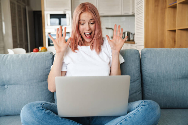 Lovely young attractive woman relaxing on a couch in the living room, using laptop computer, celebrating success - Foto, Imagem