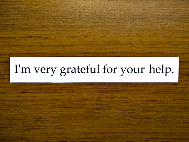 Lauseen "Olen hyvin kiitollinen avustasi". valkoisessa kirjassa, jossa on puinen kuvio tausta. - Valokuva, kuva