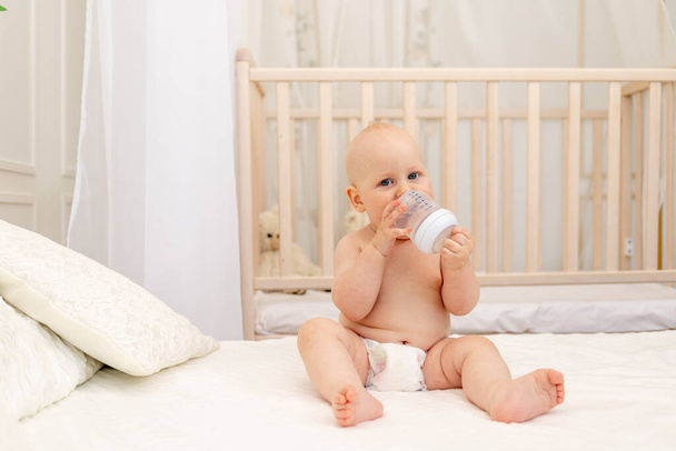 baby boy 8 months old sitting in diapers on a white bed with a bottle of milk at home, baby food concept, first bait, place for text - Foto, imagen