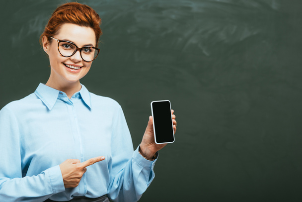 happy teacher in eyeglasses pointing at smartphone with blank screen near chalkboard  - Foto, afbeelding