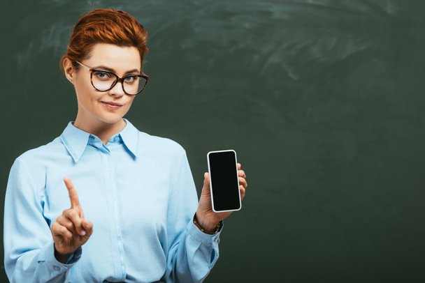 skeptical teacher showing forbidding gesture while holding smartphone with blank screen near chalkboard - Foto, Imagen