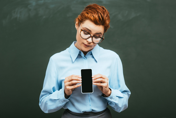 mooie jonge leraar staan in de buurt van schoolbord en het houden van smartphone met blanco scherm - Foto, afbeelding