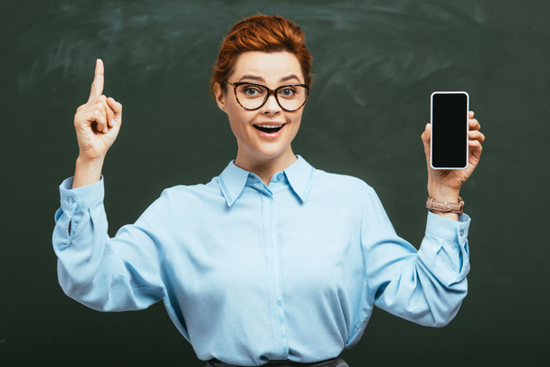 cheerful teacher in eyeglasses holding smartphone with blank screen near chalkboard    - Foto, afbeelding