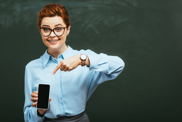 happy, attractive teacher pointing with finger at smartphone with blank screen near chalkboard  - Foto, Imagem