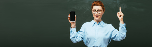 concepto horizontal de profesor sonriente mostrando gesto de idea mientras sostiene el teléfono inteligente con pantalla en blanco cerca de pizarra
 - Foto, imagen