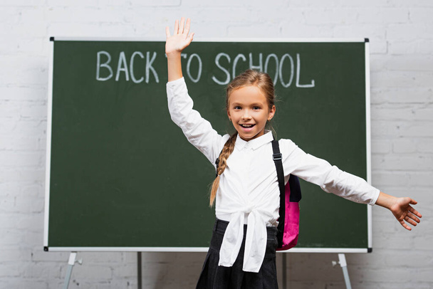 selective focus of happy schoolgirl with backpack and open arms smiling at camera near chalkboard with back to school lettering  - Foto, afbeelding