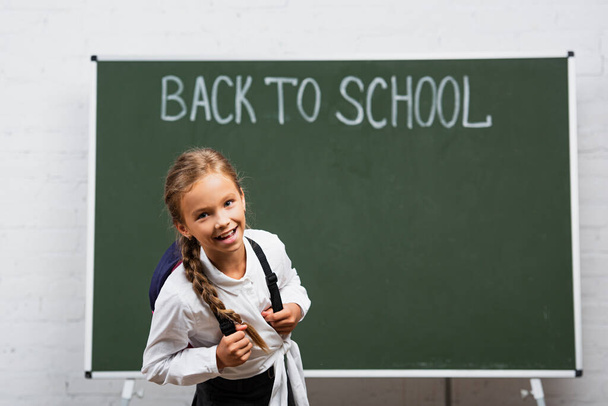 cheerful schoolgirl with backpack smiling at camera near chalkboard with back to school inscription - Foto, Imagem