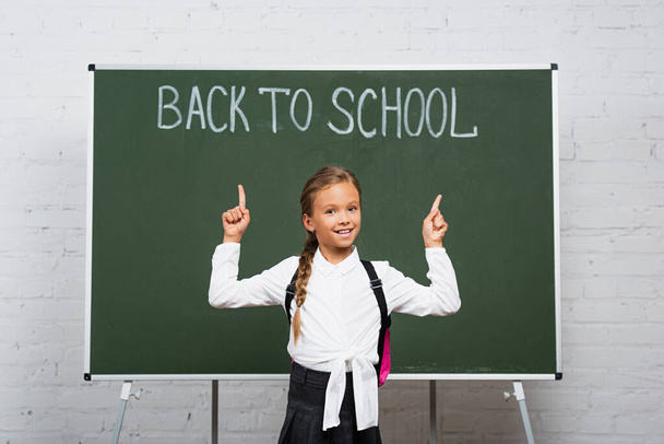 cheerful schoolgirl pointing with fingers at back to school lettering on chalkboard - Foto, immagini
