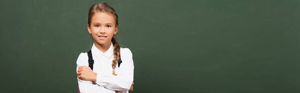 Panoramaaufnahme eines lächelnden Schulmädchens, das mit verschränkten Armen neben der Tafel steht und in die Kamera blickt - Foto, Bild