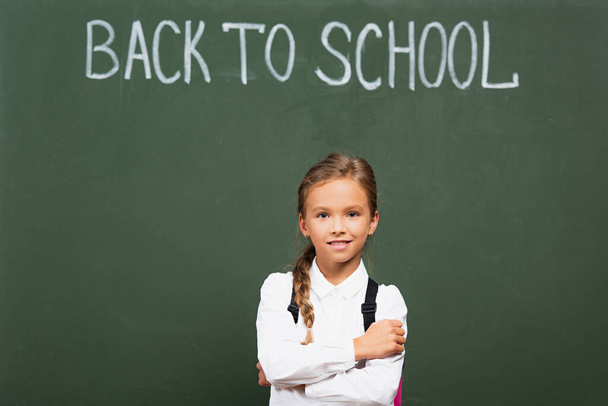 mosolygó iskolás lány áll keresztbe tett karokkal közel táblán vissza az iskolába felirat - Fotó, kép