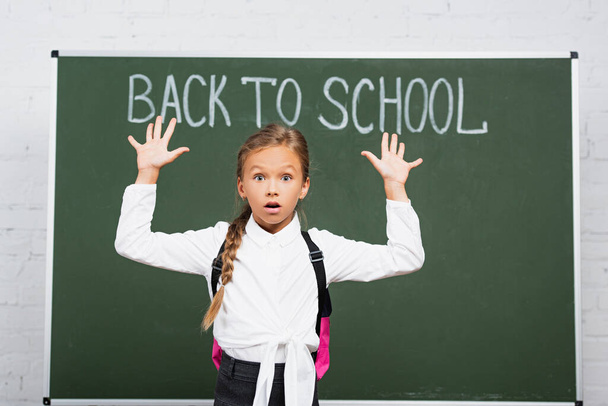 shocked schoolgirl standing with raised hands near chalkboard with back to school inscription - Foto, afbeelding